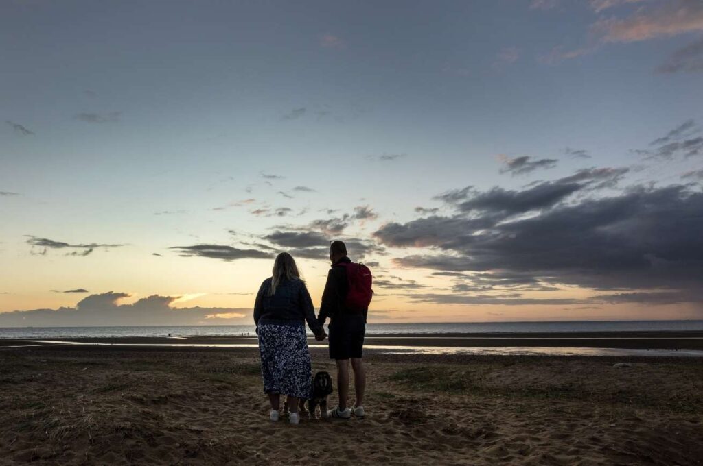 A couple hold hands and watch the sunset from a beach in Norfolk