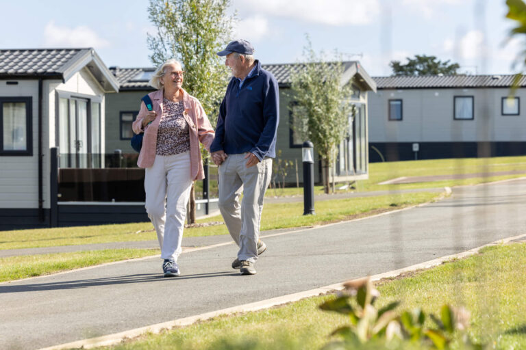 A retired couple walk hand in hand down a path through a caravan park, smiling