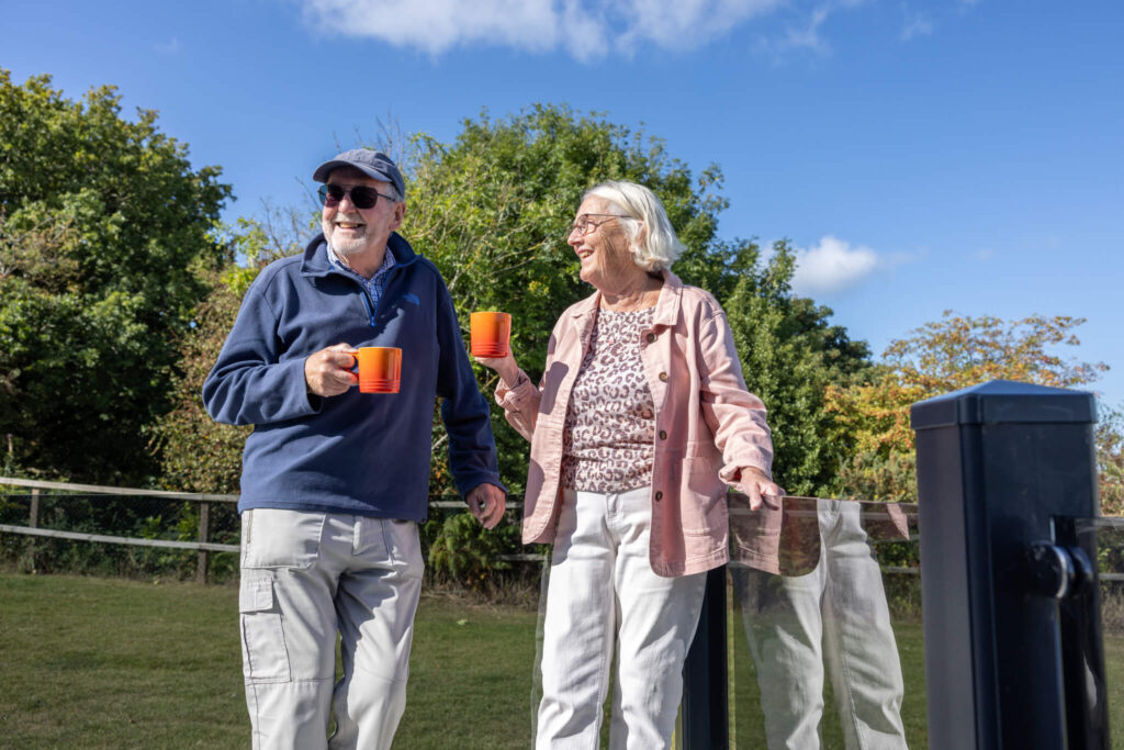 A couple enjoy a cup of tea from orange mugs on the decking of a caravan on a sunny day