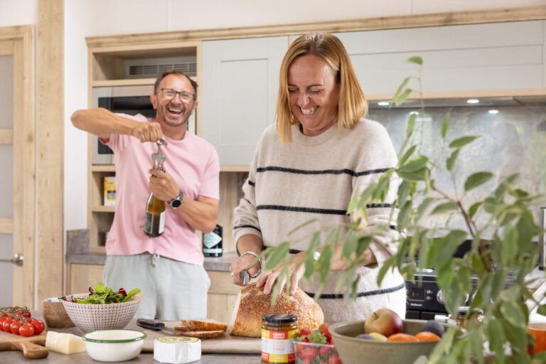 A man opens a bottle of wine while his partner cuts bread, laughing