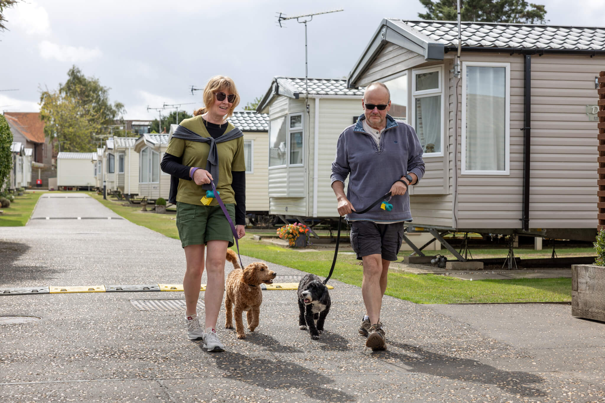 A couple wearing shorts walk their two dogs in a caravan park