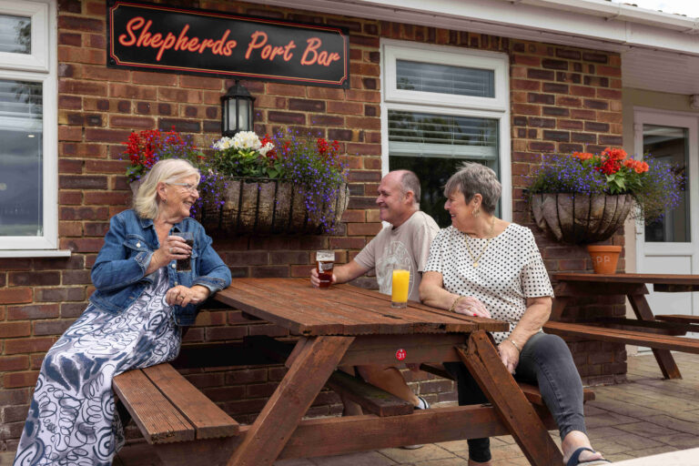 Two women and a man with drinks laugh and chat by Shepherds Port Bar